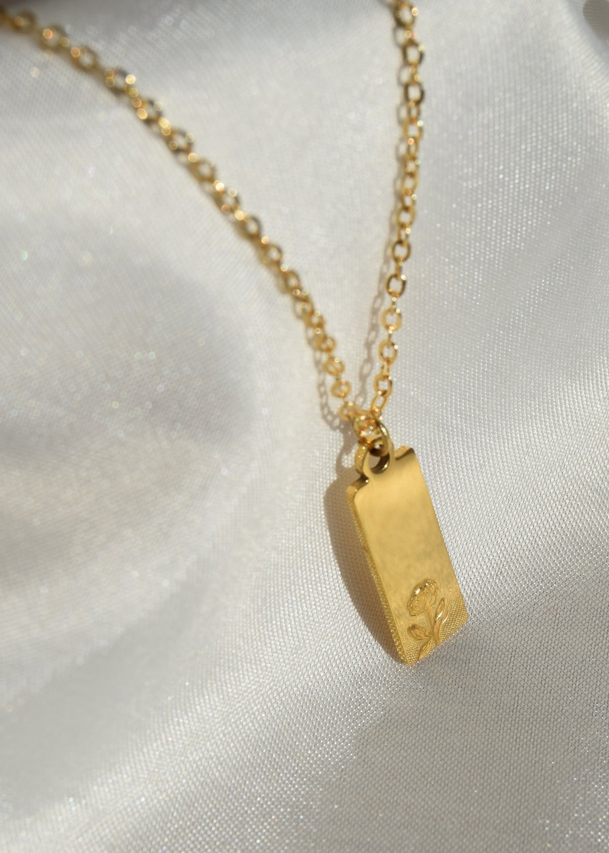 A gold chain necklace with a small rectangular gold pendant featuring an engraved clover, displayed on a textured white satin fabric.