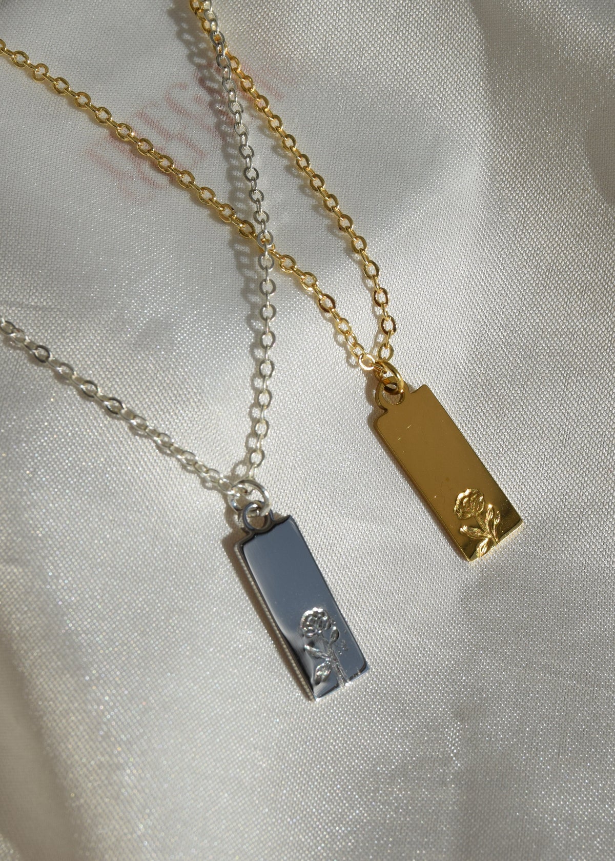 Two rectangular pendant necklaces with engraved rose details, one in gold and one in silver, displayed on draped white satin fabric.