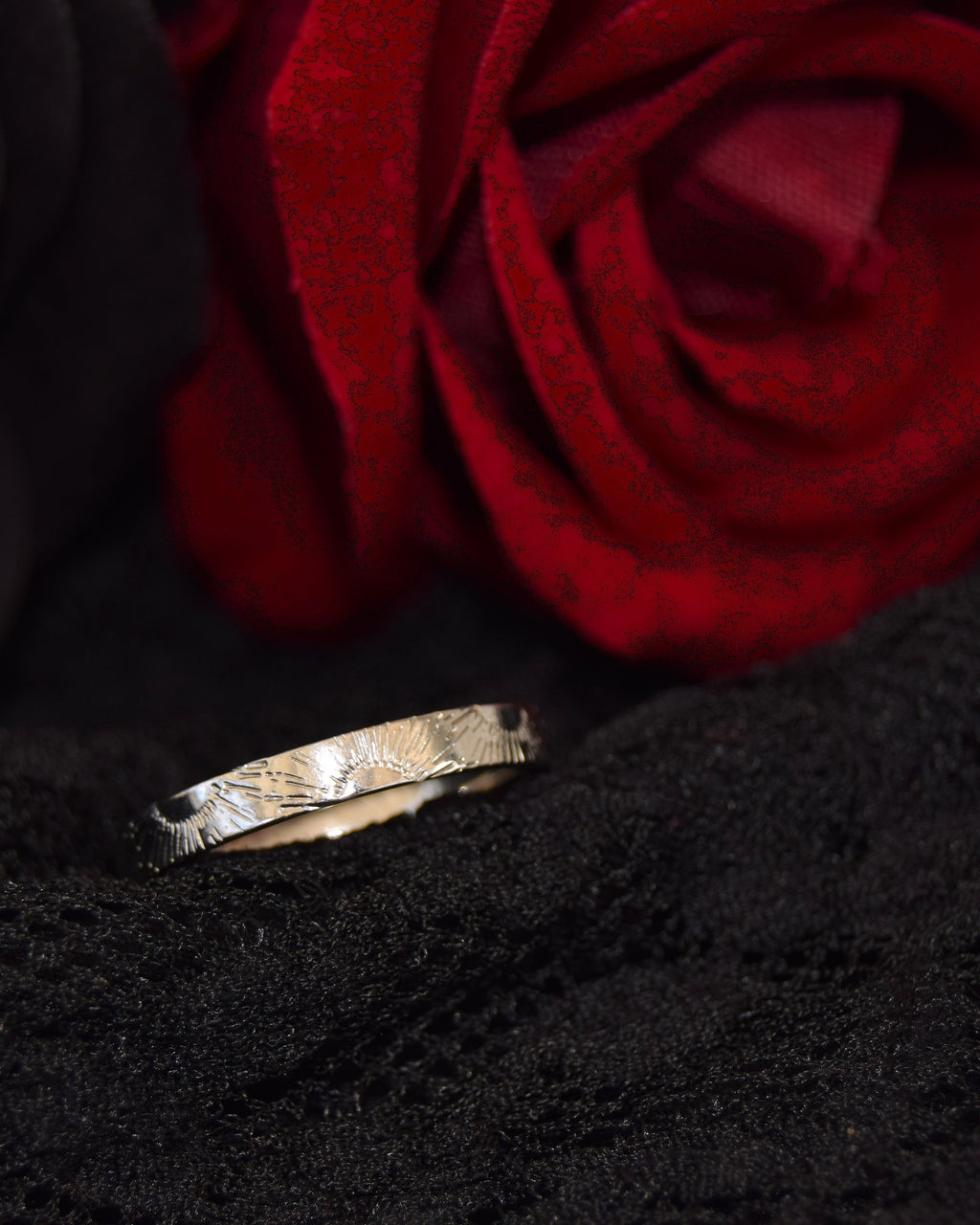 A textured silver band ring resting on a black fabric surface, with a blurred black rose on the left and a blurred red rose on the right.