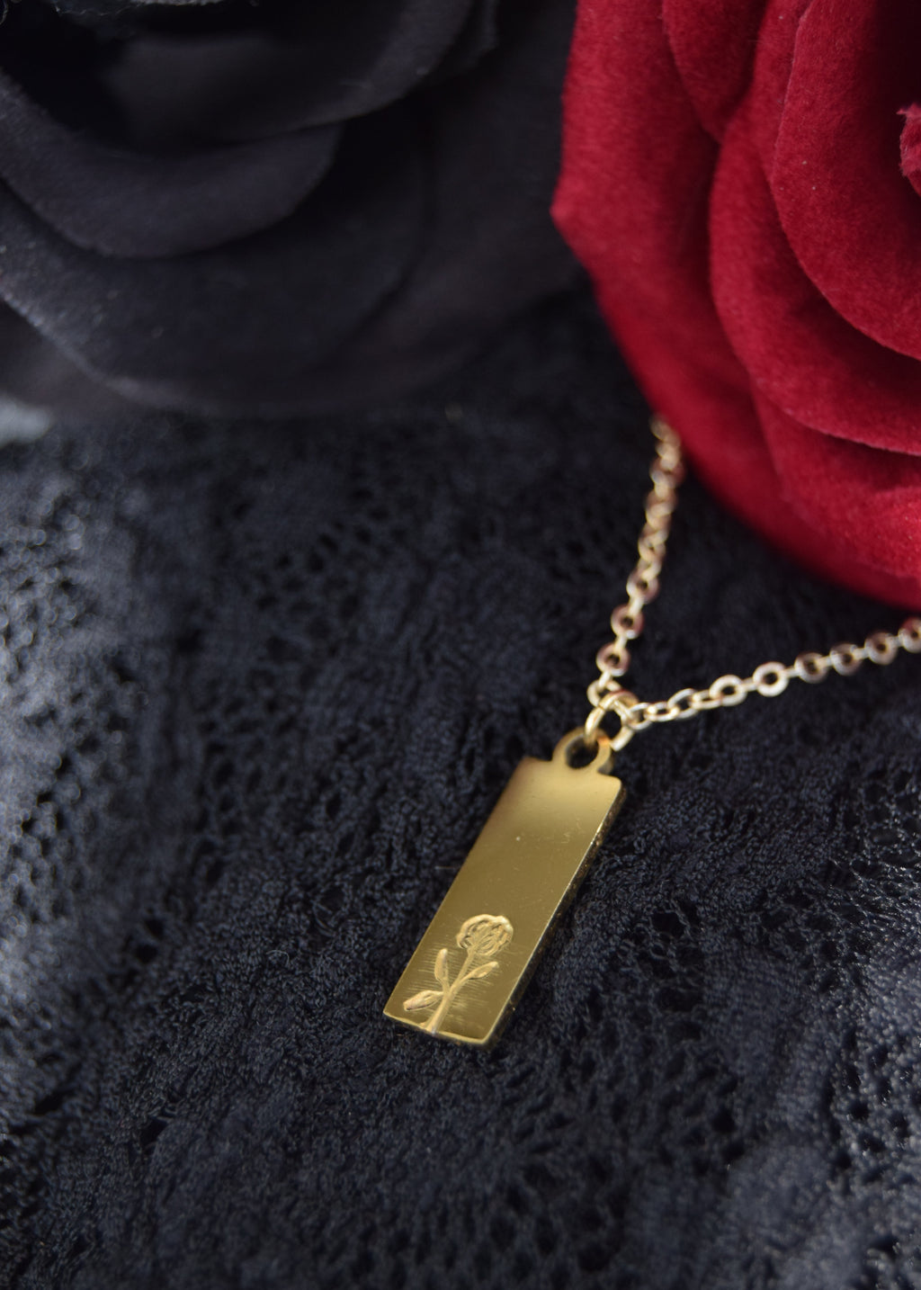 A gold rectangular pendant necklace engraved with a rose, resting on a black lace fabric next to a red rose.