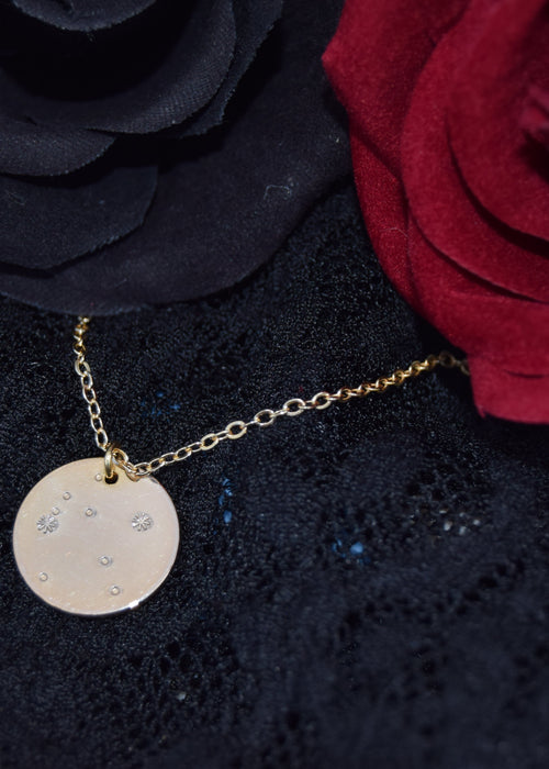 A gold-toned chain necklace with a round disc pendant featuring a constellation design and tiny rhinestones, displayed on a textured black lace surface with black and red roses.